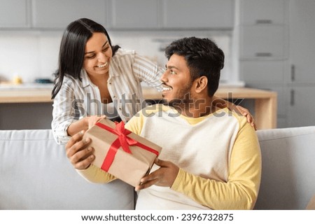 Loving indian wife surprising her husband with gift at home, greeting him with birthday or anniversary, happy eastern woman giving present box to excited man, celebrating valentine's day together Royalty-Free Stock Photo #2396732875