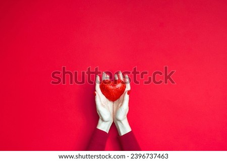 The hands of a white woman in red holding little red sparkling heart over red background