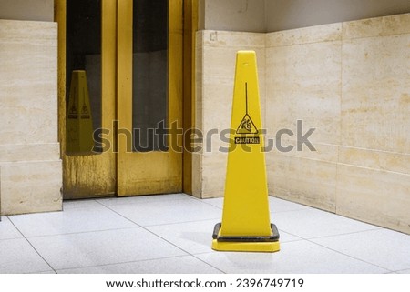 A bright yellow caution sign warns of potential hazards, standing out against the marble backdrop and reflective elevator doors