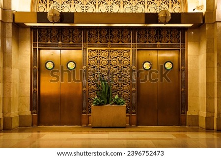 Elegant bronze elevator doors with intricate metalwork and art deco patterns, flanked by marble walls and lush greenery in a planter.