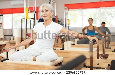 Senior woman visitor of fitness club is engaged in Pilates simulator. Improvement of well-being, physical condition, development of attractive body and figure during lessons at reformer. Royalty-Free Stock Photo #2396810719