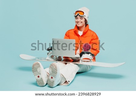 Full body fun young IT woman wear windbreaker jacket hat ski goggles mask sit with snowboarding using laptop pc computer travel rest spend winter season in mountains isolated on plain blue background