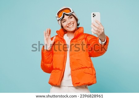 Skier young woman wearing padded windbreaker jacket ski goggles mask do selfie shot mobile cell phone wave hand travel rest spend weekend winter season in mountains isolated on plain blue background