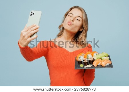 Young woman wear orange casual clothes doing selfie shot on mobile cell phone posing hold eat raw fresh sushi roll served on black plate Japanese food isolated on plain blue background studio portrait