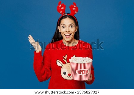 Young surprised excited merry Latin woman wear red Christmas sweater posing watch movie film hold bucket of popcorn in cinema isolated on plain blue background Happy New Year Christmas holiday concept