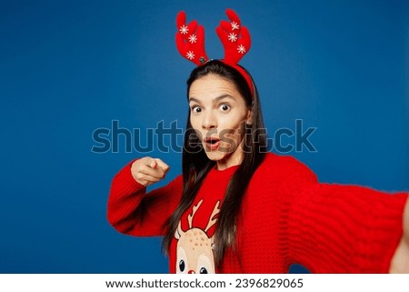 Close up young woman wear red Christmas sweater decorative deer horns on head posing do selfie shot on mobile cell phone point camera on you isolated on plain blue background. Happy New Year concept