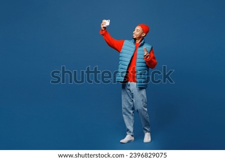 Full body young man of African American ethnicity wear padded vest red hat do selfie shot on mobile cell phone show v-sign isolated on plain dark royal navy blue background studio. Lifestyle concept
