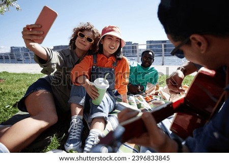 Multiethnic group of young people taking selfies outdoors enjoying picnic on grass in sunlight, copy space