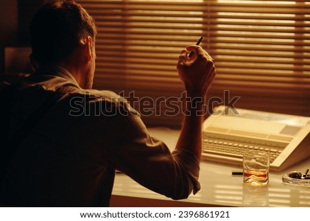 Rear view of young male clerk or police officer with cigarette in hand sitting by workplace in dark office and concentrating on work Royalty-Free Stock Photo #2396861921
