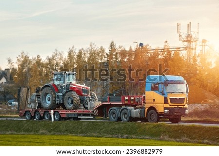 The semi truck is hauling farm equipment on a flatbed. The heavy industrial truck semi trailer flatbed platform transport one big modern farming tractor machine on the road. Evening sun. Royalty-Free Stock Photo #2396882979