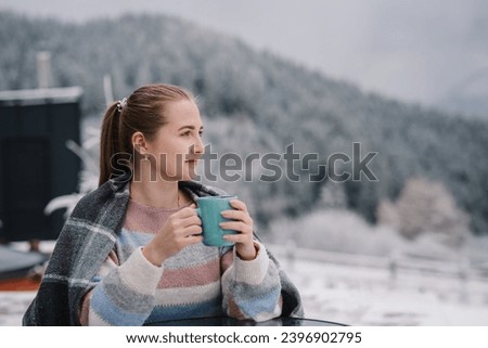 A woman in a blanket in winter looking on hill near a forest. Female sitting and holding a cup and drinking hot coffee on winter holiday. Girl drink tea and relax on cabin terrace with a mountain view