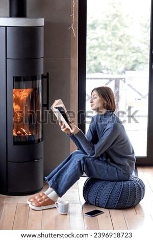 Young attractive woman sitting close to fireplace, reading interesting book and drinking hot tea in the cozy house