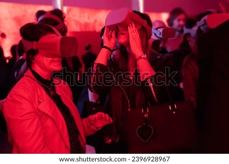 Two women immersed in virtual worlds, surrounded by a red-lit ambiance and a crowd of attendees, sharing the joy of futuristic technology at a dynamic and immersive event Royalty-Free Stock Photo #2396928967
