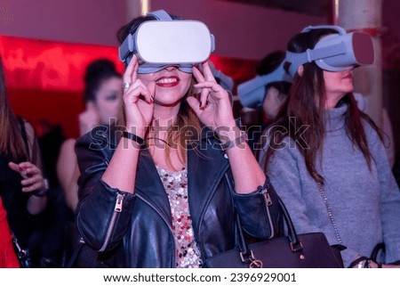 In a sea of red-lit anticipation, two women with VR glasses share smiles, surrounded by a vibrant crowd, embracing the future in a dynamic and immersive event Royalty-Free Stock Photo #2396929001