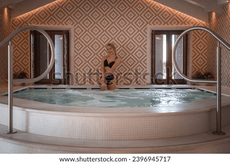 Attractive woman in black bikini standing in hot tub with tiled floor and decorative lighting. Side angle view. No steam or bubbles. Unknown location.