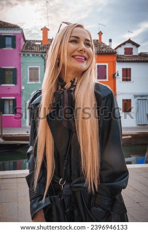 Blonde woman in black trench coat standing in front of colorful buildings in Venice, Italy. Unknown angle, expression, and architectural style. No visible signage or writing.