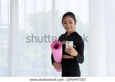 A smiling Asian woman holds a rolled-up yoga mat and takes a selfie in a yoga studio. She is wearing sporty clothing and looks happy and healthy. 