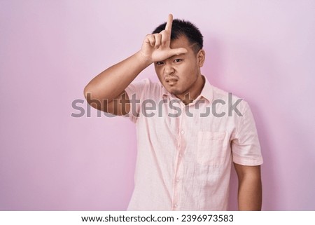 Chinese young man standing over pink background making fun of people with fingers on forehead doing loser gesture mocking and insulting. 