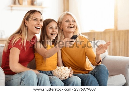 Positive three generations of women family watching TV together at home, happy mother, teen daughter and grandmother sitting on couch with remote control and bowl with popcorn, copy space