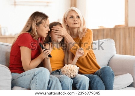 Family three generations of women watching TV together at home, grandmother, mother and grandchild teen girl sitting on couch, eating popcorn, watching horror, adult women covering child eyes