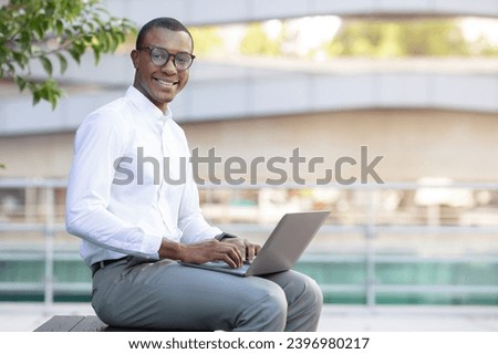 Portrait Of Handsome Black Businessman In Eyeglasses Working On Laptop Outdoors, Smiling African American Male Entrepreneur Using Computer Outside, Sitting On Bench Near Modern Office Building
