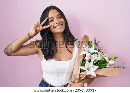 Brunette woman holding bouquet of white flowers doing peace symbol with fingers over face, smiling cheerful showing victory 
