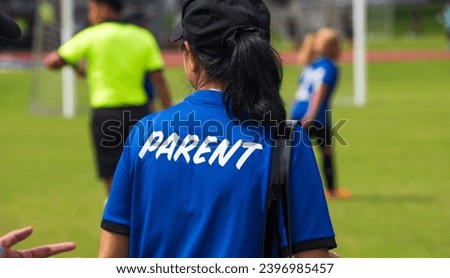 Mom standing and watching her son playing football in a school football tournament on a sideline with a sunny day. Sport, outdoor active, lifestyle, happy family and soccer mom and soccer dad concept. Royalty-Free Stock Photo #2396985457