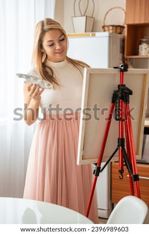 Beautiful young blonde woman artist with palette and brush painting abstract pink picture on canvas at home. Art and creativity concept. High quality photo