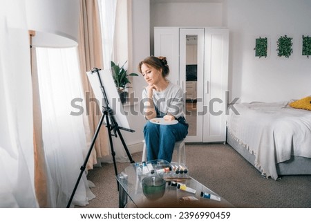 female artist sitting in front of easel, painting picture with inspiration, drawing on canvas with paintbrush, palette, smiling, relaxing at home. Art, hobby, leisure and creative occupation concept