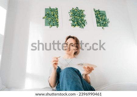 Smiling female artist sitting on bed with paintbrush, palette under her hand-drawn painting art works on the wall at home. Art, hobby, leisure and creative occupation concept. DIY, Home decorating.