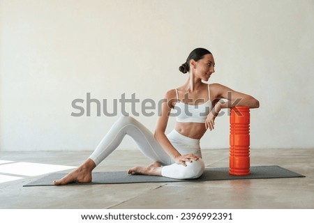 With orange colored roller. Fitness woman is indoors in white clothes. Royalty-Free Stock Photo #2396992391