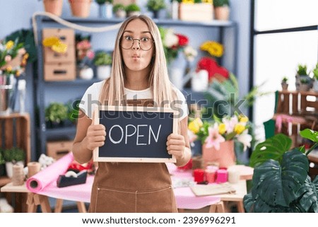 Young blonde woman working at florist holding open sign making fish face with mouth and squinting eyes, crazy and comical. 