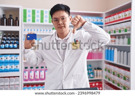 Young asian man working at pharmacy drugstore holding credit card with angry face, negative sign showing dislike with thumbs down, rejection concept 