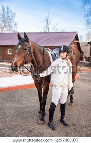 Pretty blond professional female jockey standing near brown horse on farm in winter. Friendship with horse concept. High quality photo
