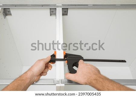 Young adult man hands using f speed clamp and pressing white boards of base cabinet frame. Assembling new kitchen wooden furniture. Renovation process. Closeup. Point of view shot. Royalty-Free Stock Photo #2397005435
