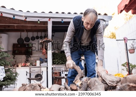 Photo of an elderly man stacking firewood in front of his house.