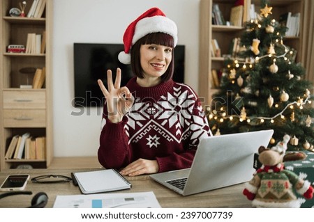 Caucasian businesswoman in Santa hat looking at camera showing sign ok sitting in office near xmas tree and working on laptop during the holiday season, typing on computer keyboard.