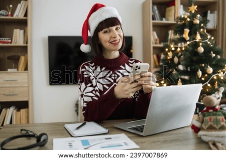 Young adult female freelance worker in Santa hat holding smartphone posing to camera while staying in workplace at home at background of Christmas tree.