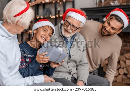 Happy multi generation family celebrating Christmas while little boy opening Christmas present. New Year Eve and winter holiday spirit atmosphere. Gift present sharing giving concept