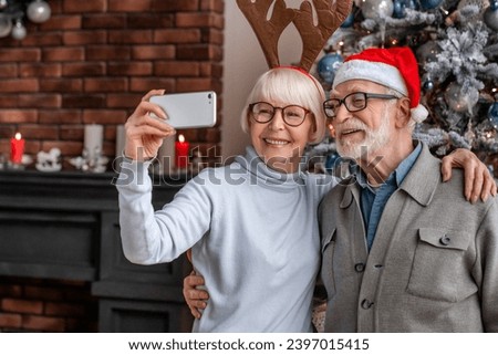 Happy senior couple taking a selfie and having fun at home while celebrating Christmas Eve. Elderly old grandparents spouses using smart phone for social media posting