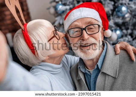 Happy smiling senior couple in hats taking selfie at home. Christmas celebration of elderly spouses grandparents vlogging blogging in front of fir tree