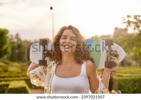 portrait of a smiling woman with curly hair holding a skateboard behind her head