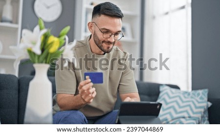 Young hispanic man shopping with touchpad and credit card sitting on sofa at home