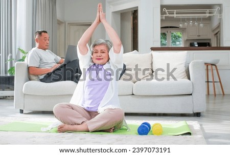 Asian aging senior old woman wearing casual comfortable t shirt, sitting on floor in living room at cozy home, doing yoga exercise with happiness, smiling. Healthcare, Retirement Concept