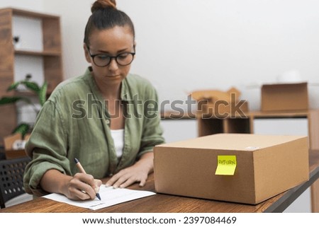 Serious manager in glasses writes application to send cardboard box with yellow sticker with goods back woman issues return to online store sitting at desk with parcel Royalty-Free Stock Photo #2397084469