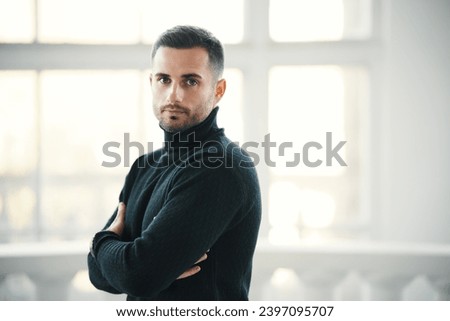 Confident handsome man in a black turtleneck stands with arms crossed in modern interior with copy space. Male beauty concept Royalty-Free Stock Photo #2397095707