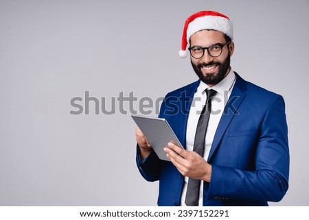Arabian middle-eastern businessman in Santa Claus red hat using digital tablet for Christmas shopping online, texting, e-commerce, remote work isolated over grey background Royalty-Free Stock Photo #2397152291