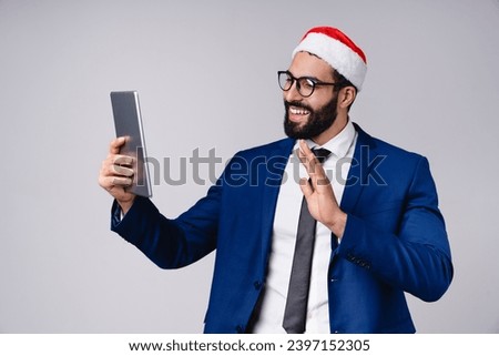 Videocall conference meeting. Arabian businessman in Santa Claus red hat using digital tablet for online shopping for Christmas New Year, texting, surfing social media on grey background