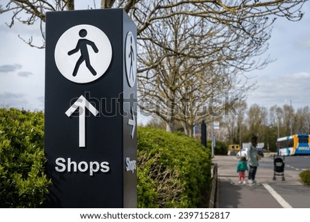 Large directional signage post with arrow and person walking icon pointing along path towards shopping retail area. Family on path in the background.