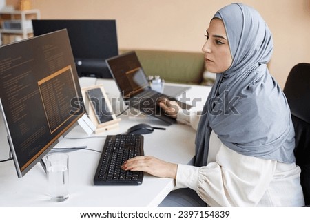 Side view portrait of Muslim young woman wearing modest clothing in office while working as female software developer and writing code with multiple computer devices